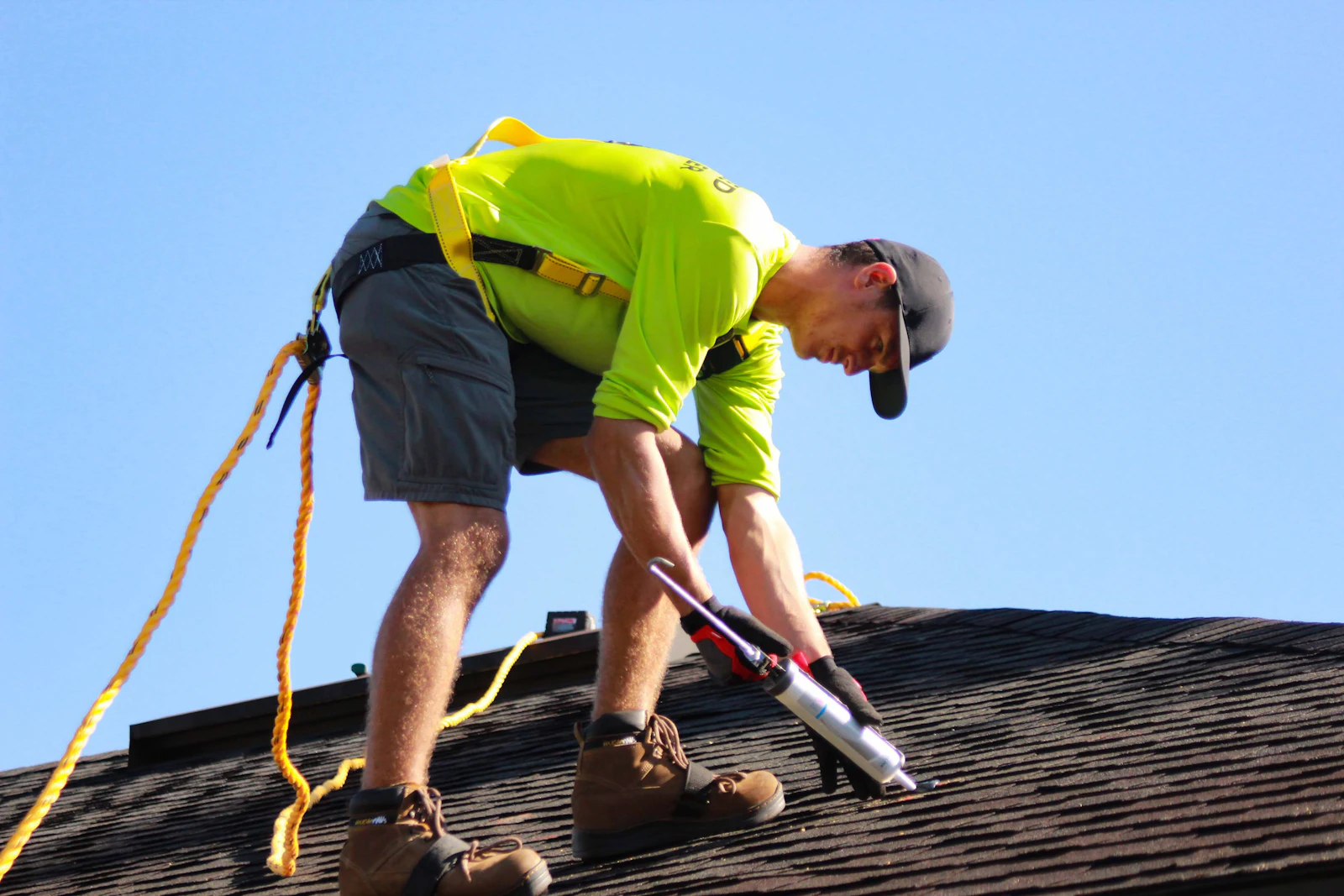 A roofer in a harness sealing a shingle roof with a caulk gun